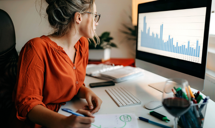 mulher branca de camisa laranja e coque analisando gráficos na tela de um computador